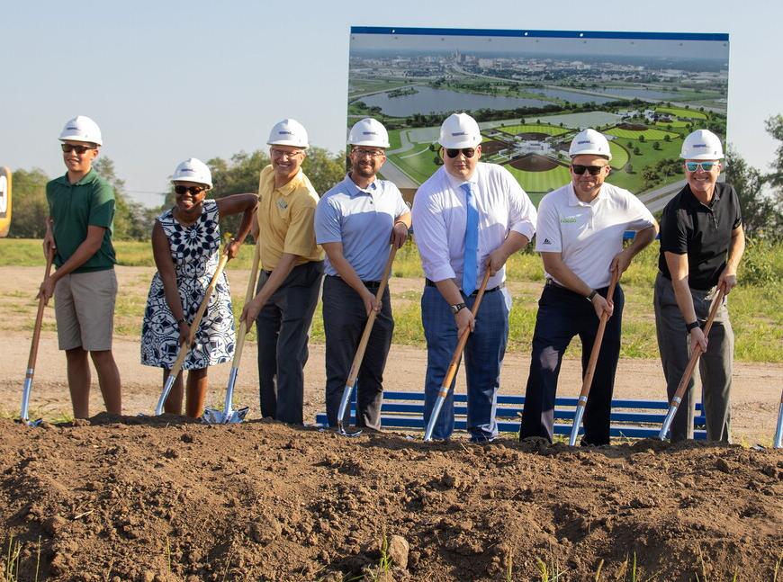 Community leaders wearing hard hats and holding shovels break ground at the new sports complex .